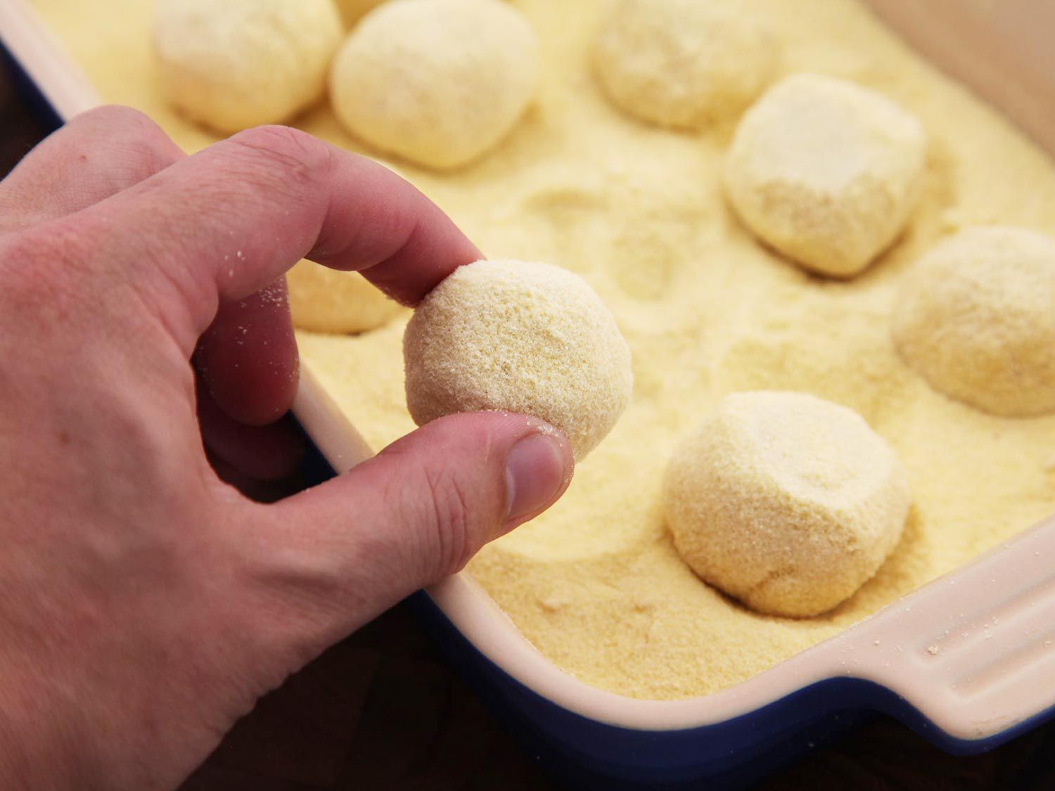 A ricotta ball between two fingers overlooking a baking dish containing gnudi in semolina. 