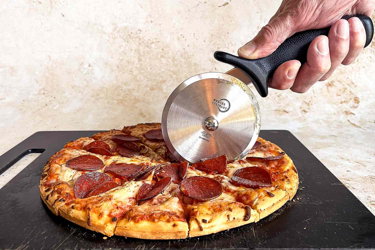 A hand using a Mercer pizza cutter to slice a pepperoni pizza