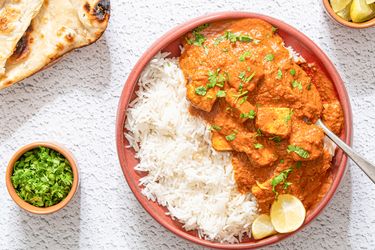 A bowl of tofu tikka masala with rice and garnished with fresh herbs accompanied by naan bread lime wedges and a small bowl of chopped herbs
