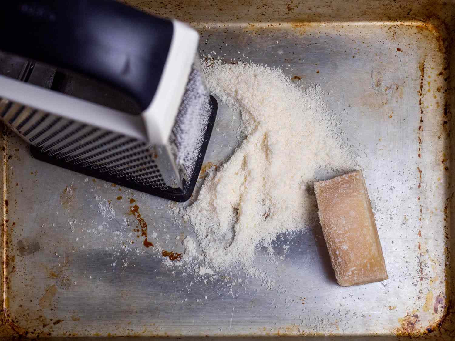 A box grater sites on a rimmed baking sheet alongside a pile of grated cheese and a chunk of Parmesan.