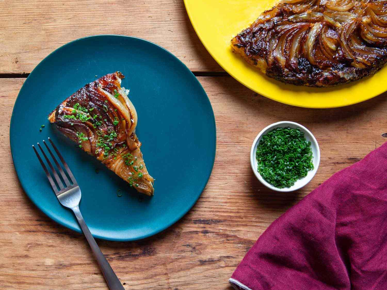 A slice of tarte tatin sprinkled with chives on a small plate, with a ramekin of chives and the rest of the tart on the side.