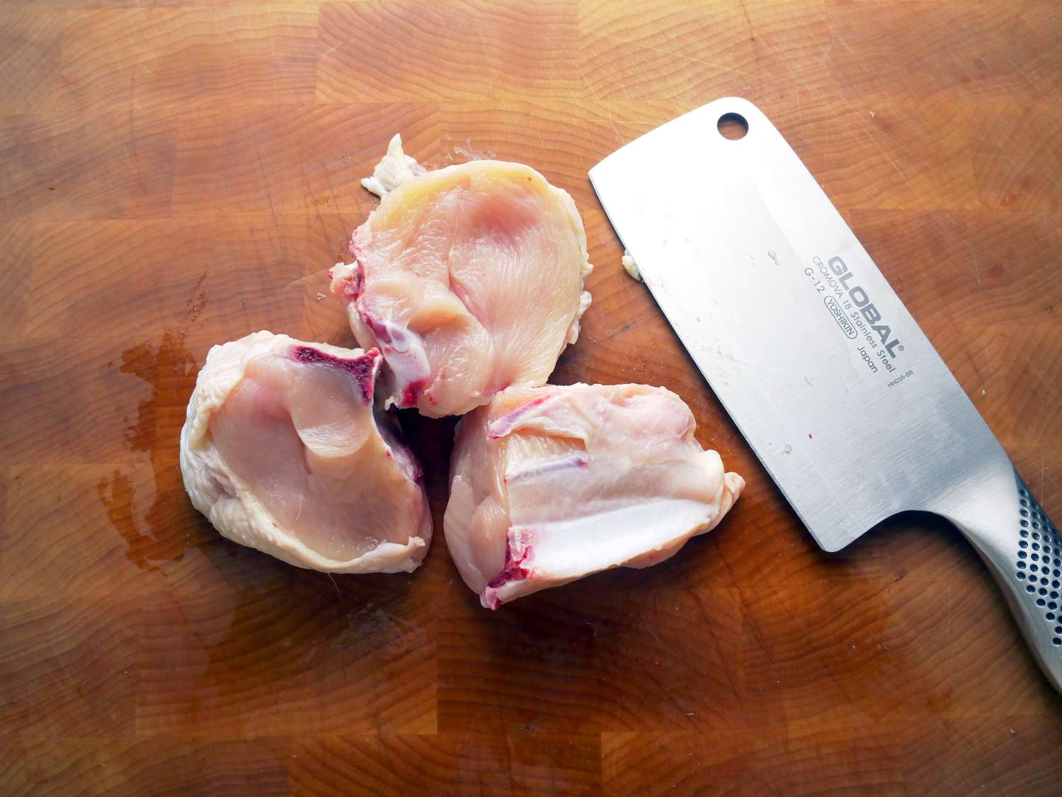 Global 6.5-Inch Meat Cleaver on a cutting board next to a bone-in chicken breast cut into thirds