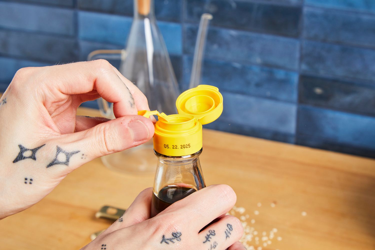 Hands opening a yellow-capped bottle with a visible expiration date, showing tattoos on fingers