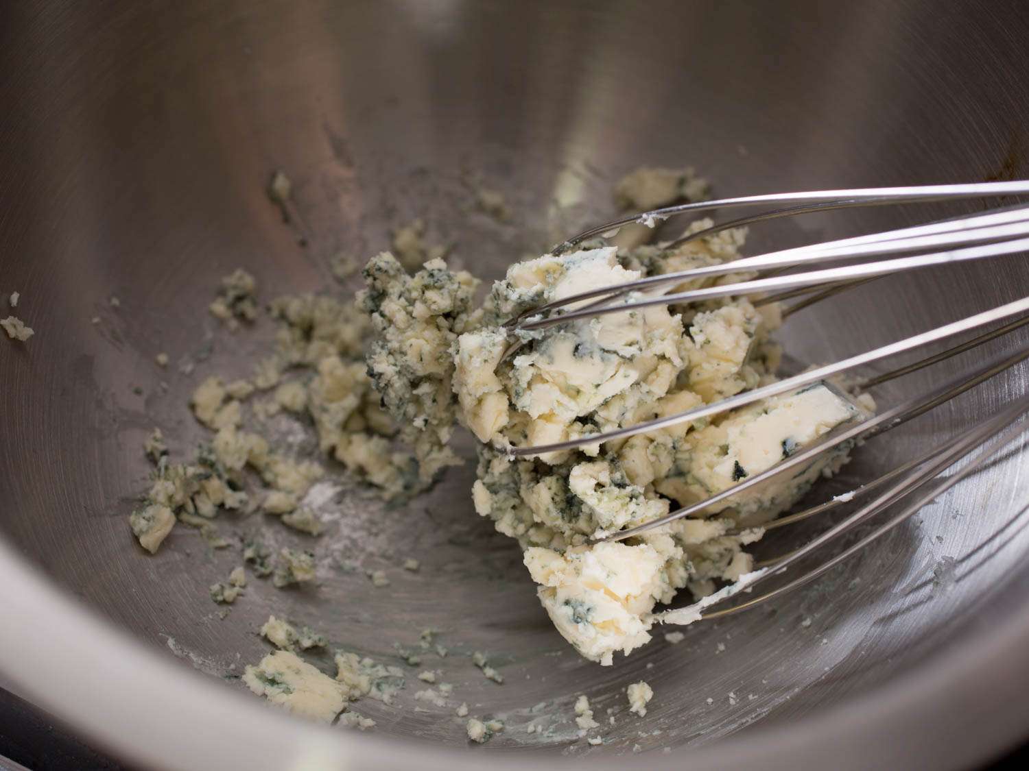 Blue cheese being broken up with a whisk in a metal bowl.
