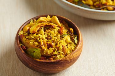 A small round wooden bowl holding poha chivda. There is a larger cream-colored ceramic bowl in the top right corner of the image.