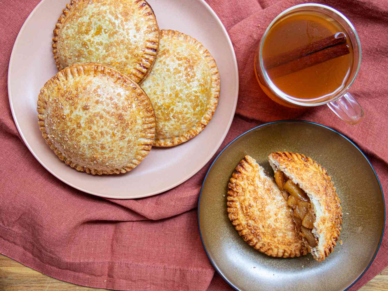 Apple Handpies on a serving bowl, with one handpie on small plate, ripped in half. Cup of warm cider, terra cotta colored textile under the dishes