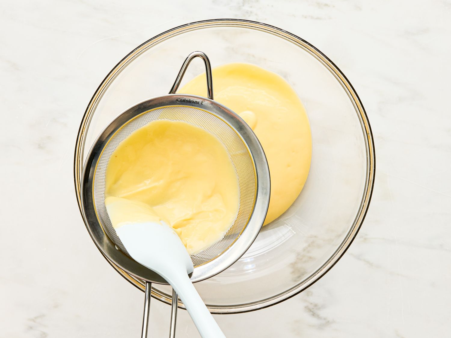 Custard being strained through a mesh sieve into a glass bowl with a spatula