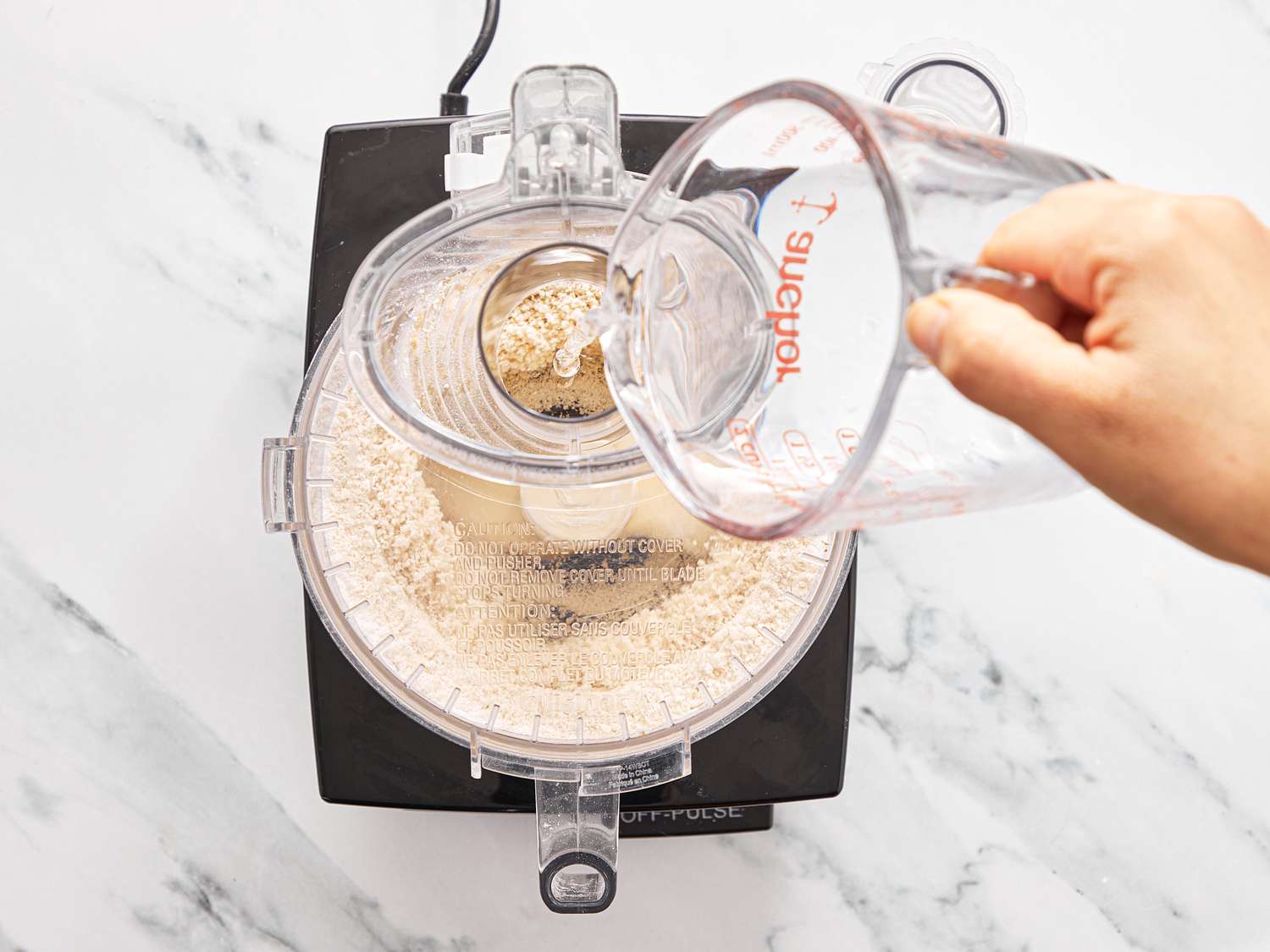 A food processor with dry ingredients being mixed and liquid being poured from a measuring cup
