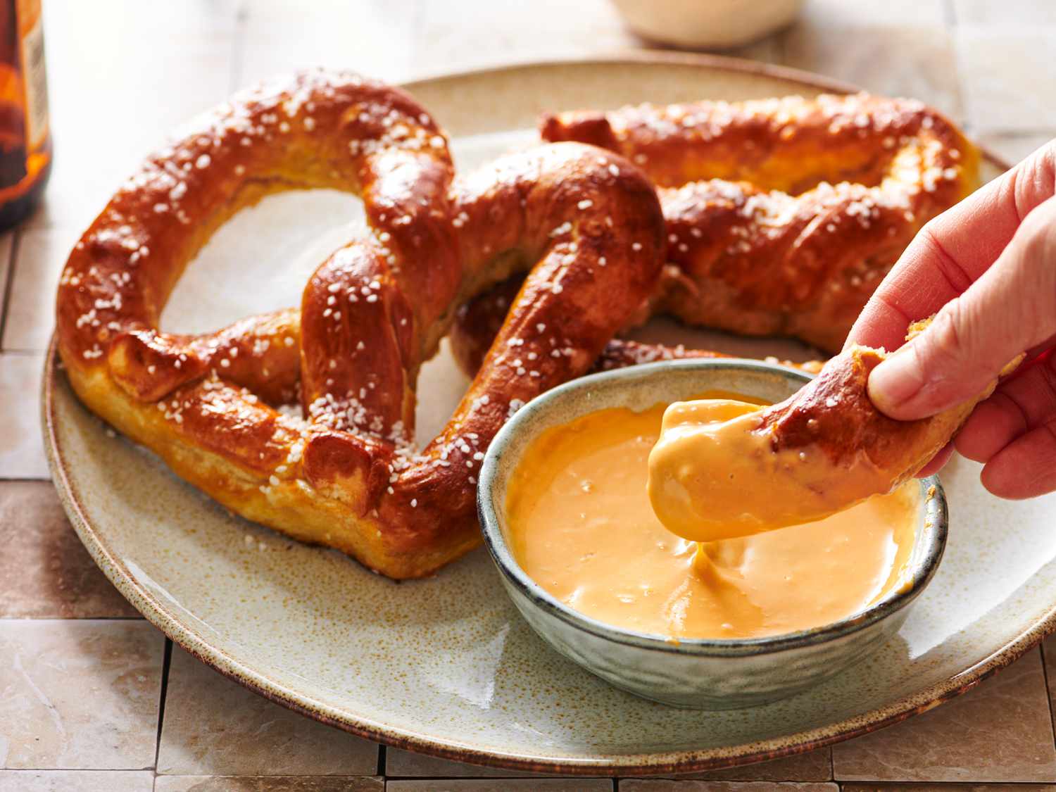 A round ceramic plate holding two Bavarian-style pretzels and a small bowl of beer cheese. Part of one pretzel is being dipped into the beer cheese by a hand.