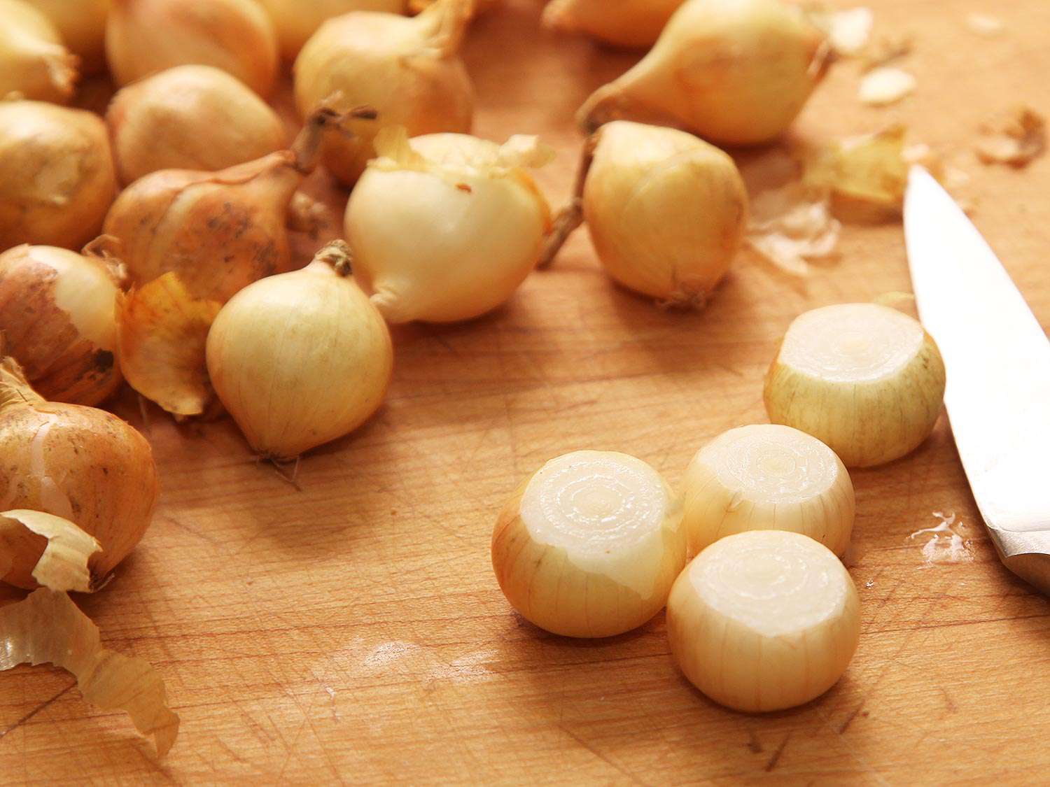 Pearl onions with their tops cut off in front of a number of pearl onions which are not yet trimmed.