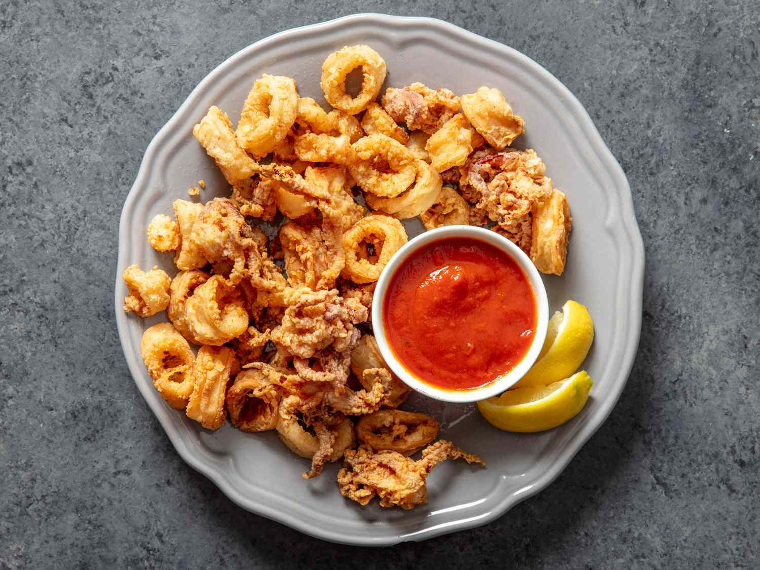 A round gray plate with a pile of fried calamari, two lemon wedges, and a small bowl of marinara sauce.