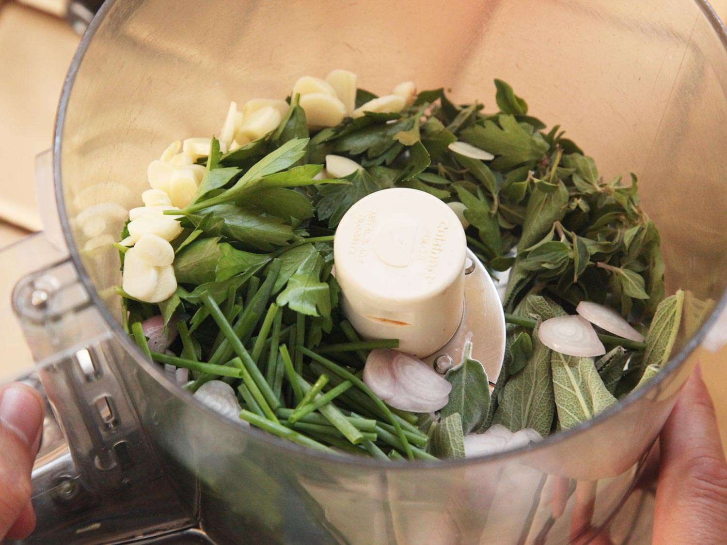 A food processor filled with unchopped green herbs, shallots, and garlic cloves.