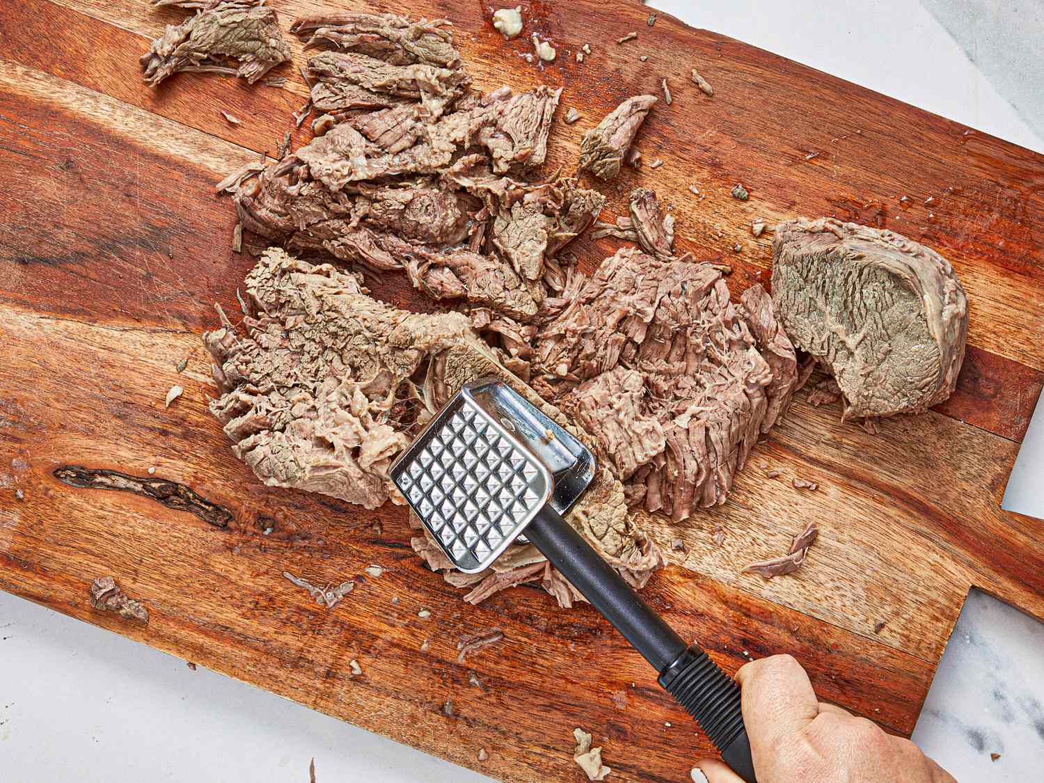 Cooked beef being tenderized with a meat mallet on a wooden cutting board