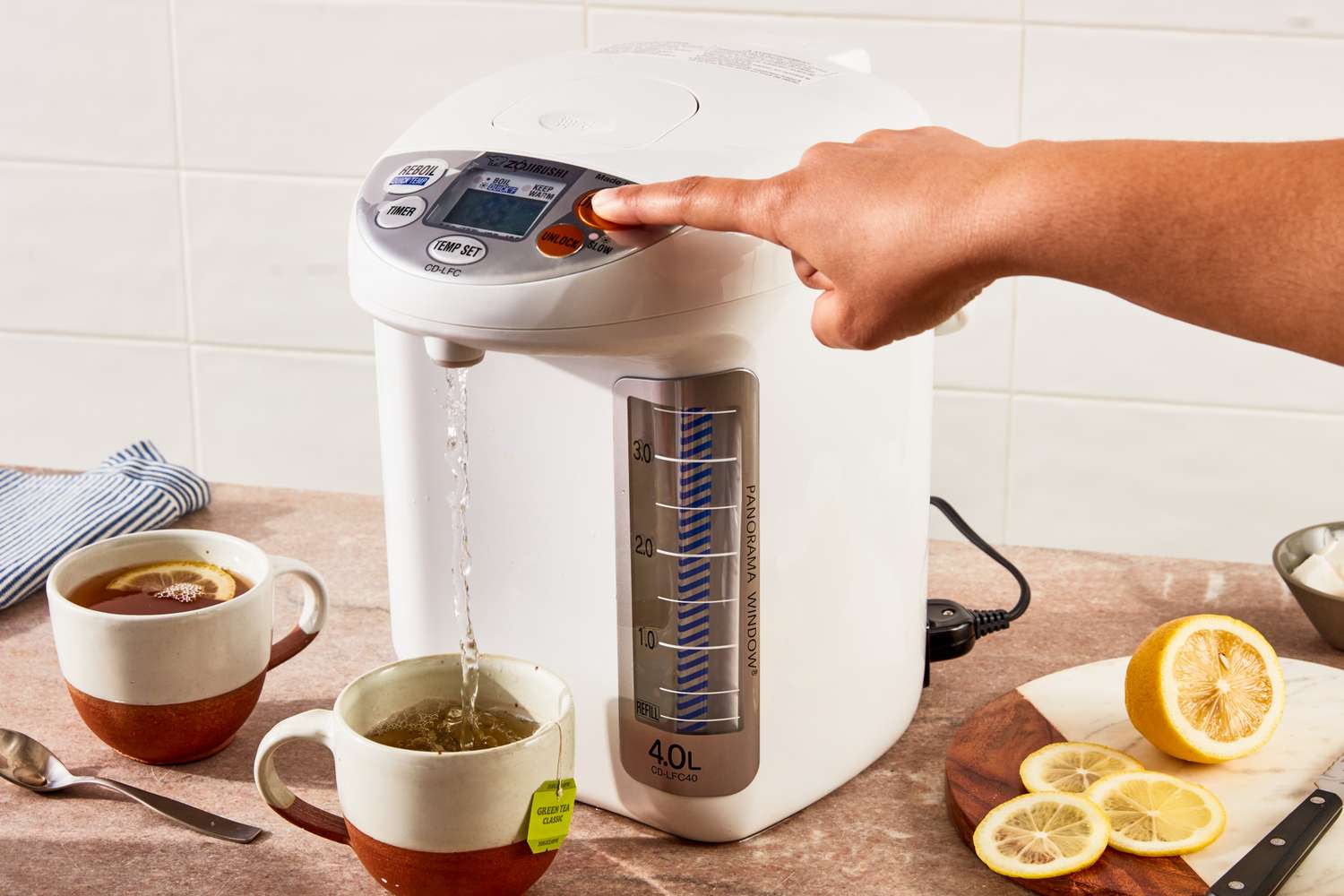A hand pressing a button on an electric water boiler while hot water fills a mug surrounded by tea and sliced lemon