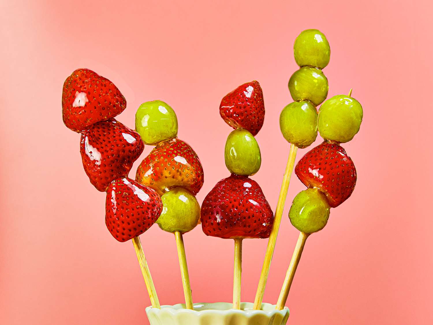 Tanghulu sticks with strawberries and grapes standing upright in cup, with a pink background