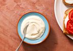 Two-minute mayonnaise in a serving bowl with a spoon, with an open face tomato sandwich on the right edge of the frame.