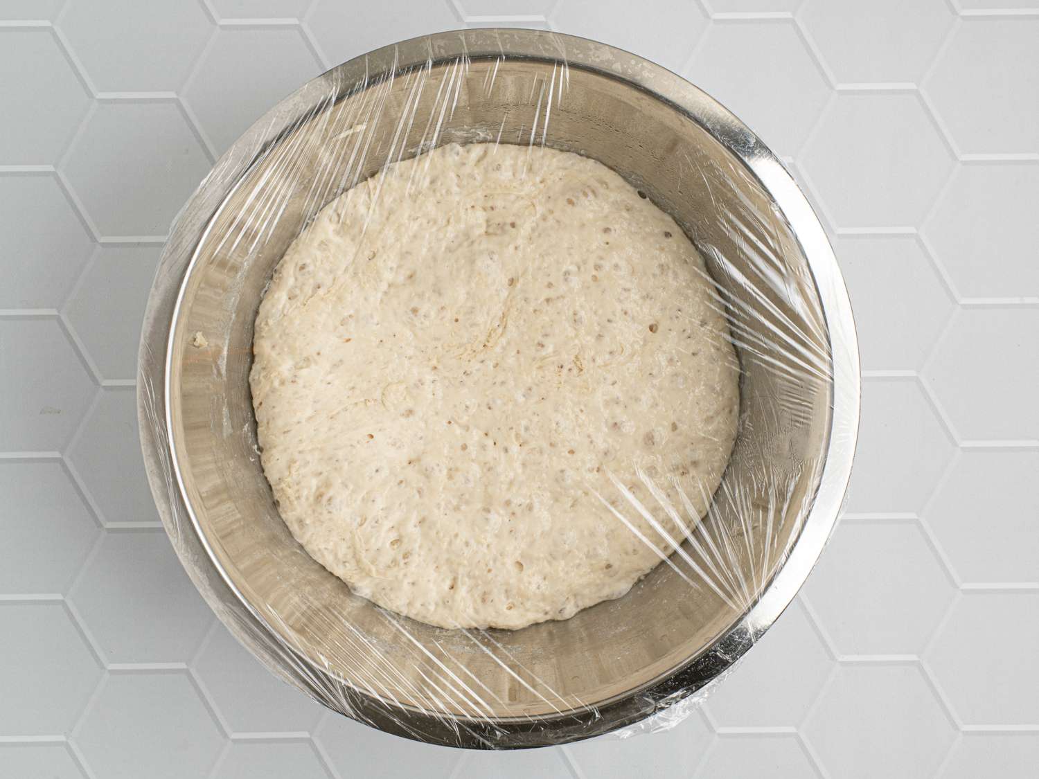 A metal bowl holding the dough, covered with plastic wrap, on a ceramic counter.