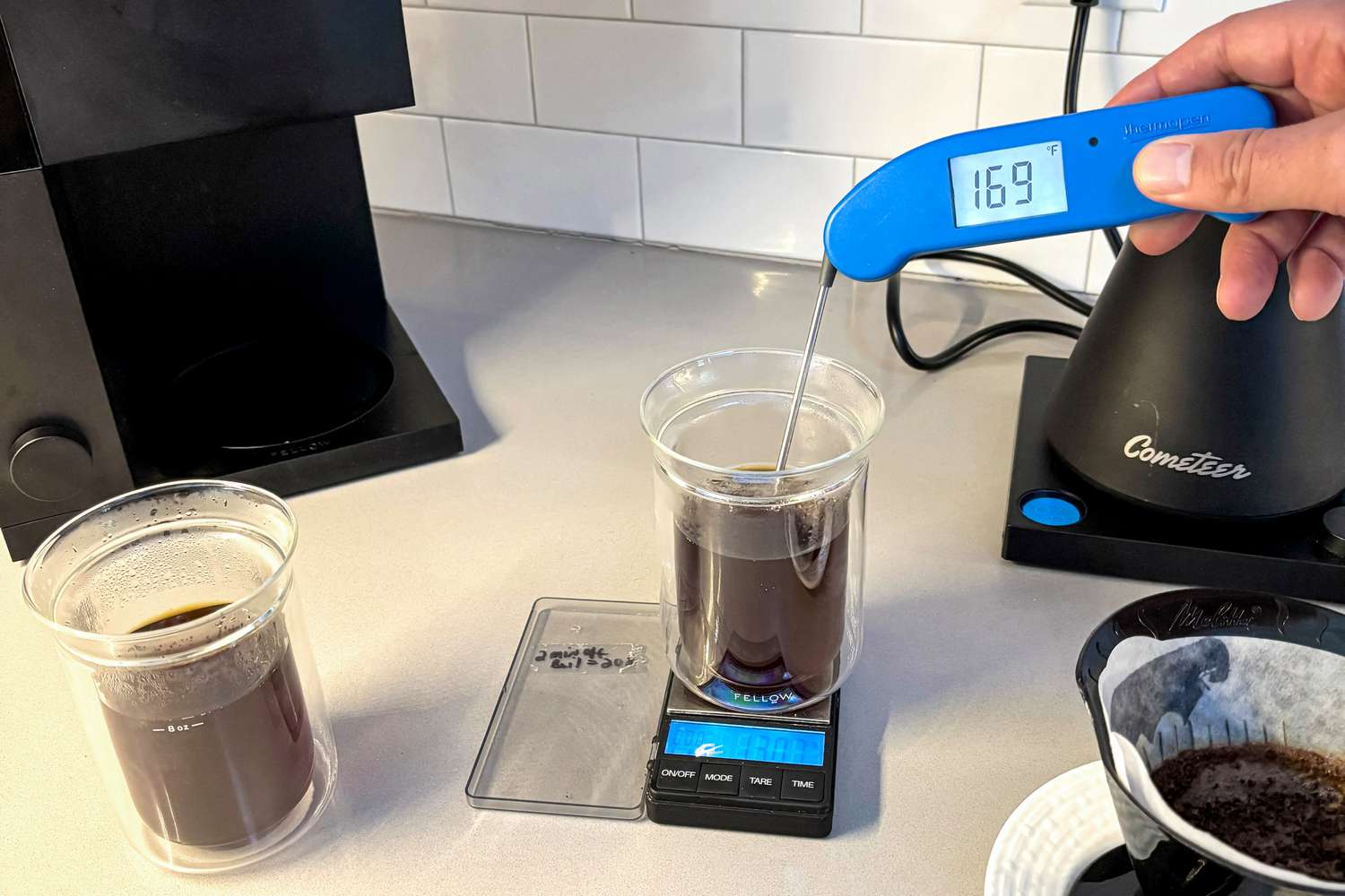 The Fellow Aiden beside a pour-over device. A person is taking the temperature of a cup of coffee.