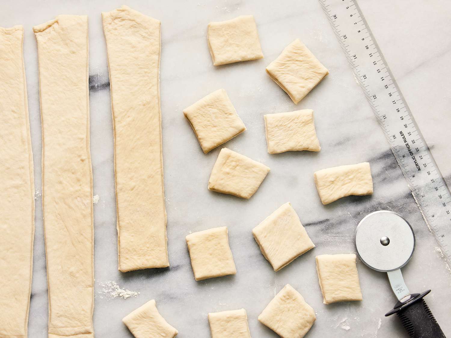 Dough being cut into strips and then small squared with a pizza cutter and ruler on marble surface 