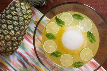 Overhead view of Isla Bonita Punch, garnished with mint leaves and lime wheels. A large sphere of ice floats in the center of the punch bowl. A whole pineapple rests on its side nearby.