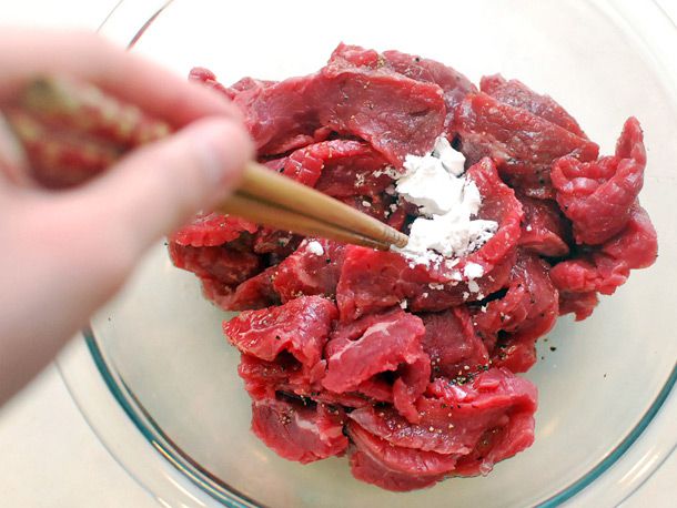Raw slices of beef in a glass bowl, topped with corn starch, with a pair of chopsticks which are about to mix the ingredients together.