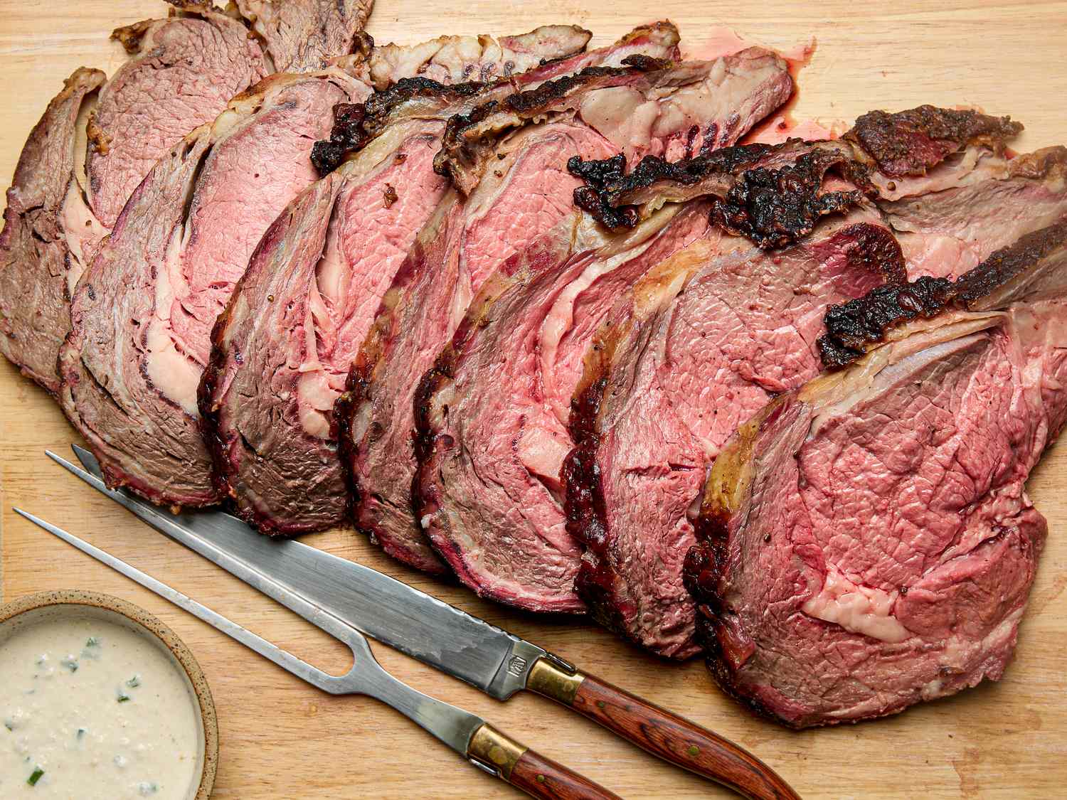Slices of cooked prime rib arranged on a cutting board with a carving knife and fork alongside a dish of sauce