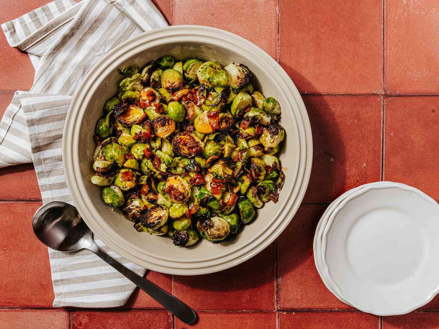 Bowl of roasted brussel sprouts on a brick surface, with a striped towl, spoon and smaller serving dishes to the side 