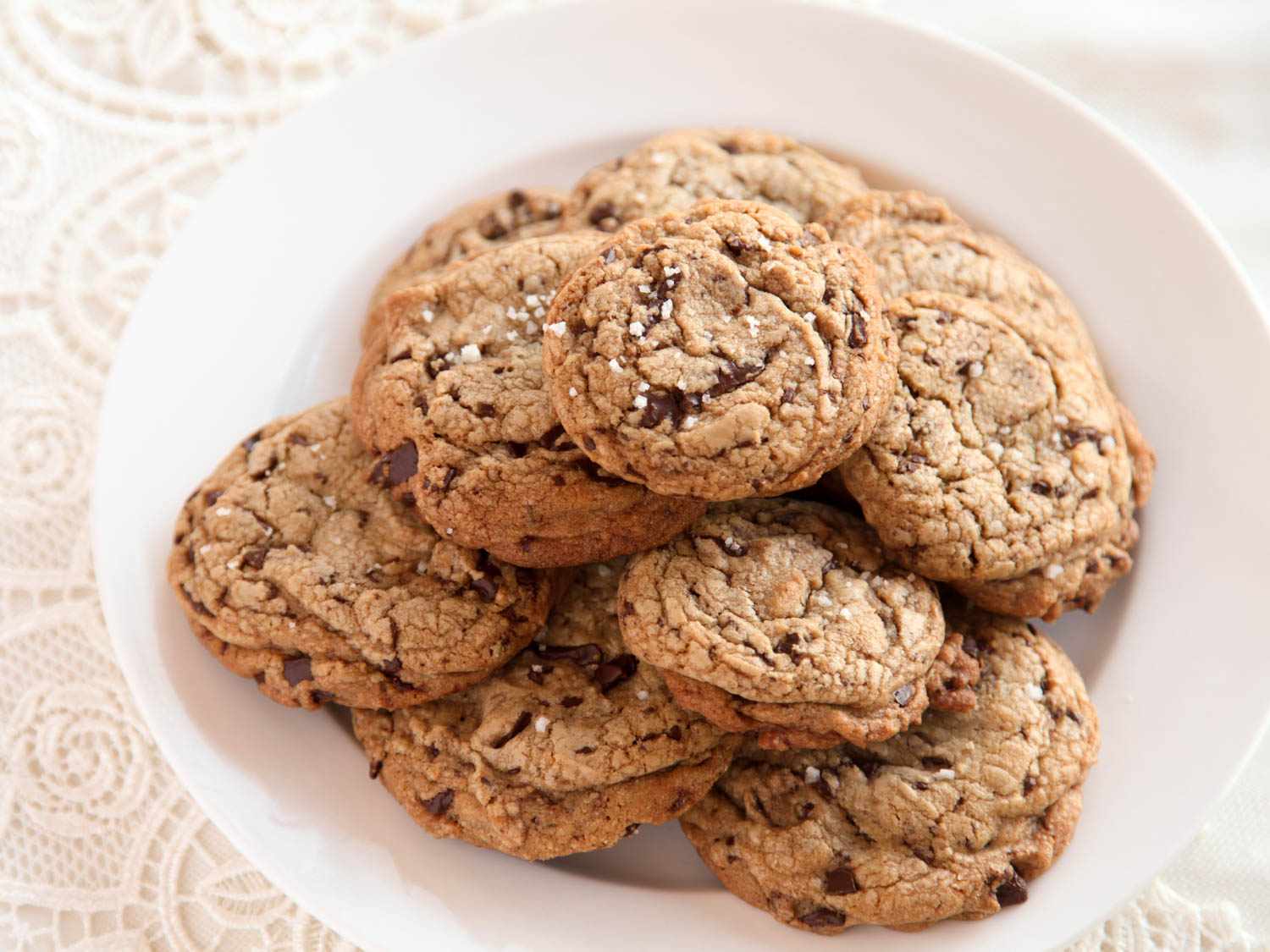 A pile of salted chocolate chip cookies on a white plate, sitting on a lace background.