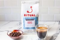 A bag of Ritual coffee labeled 'Sweet Tooth' with a bowl of coffee beans and a glass of espresso on a countertop