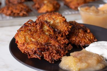 A plate of fried latkes served with applesauce and sour cream additional latkes cooling on a rack in the background