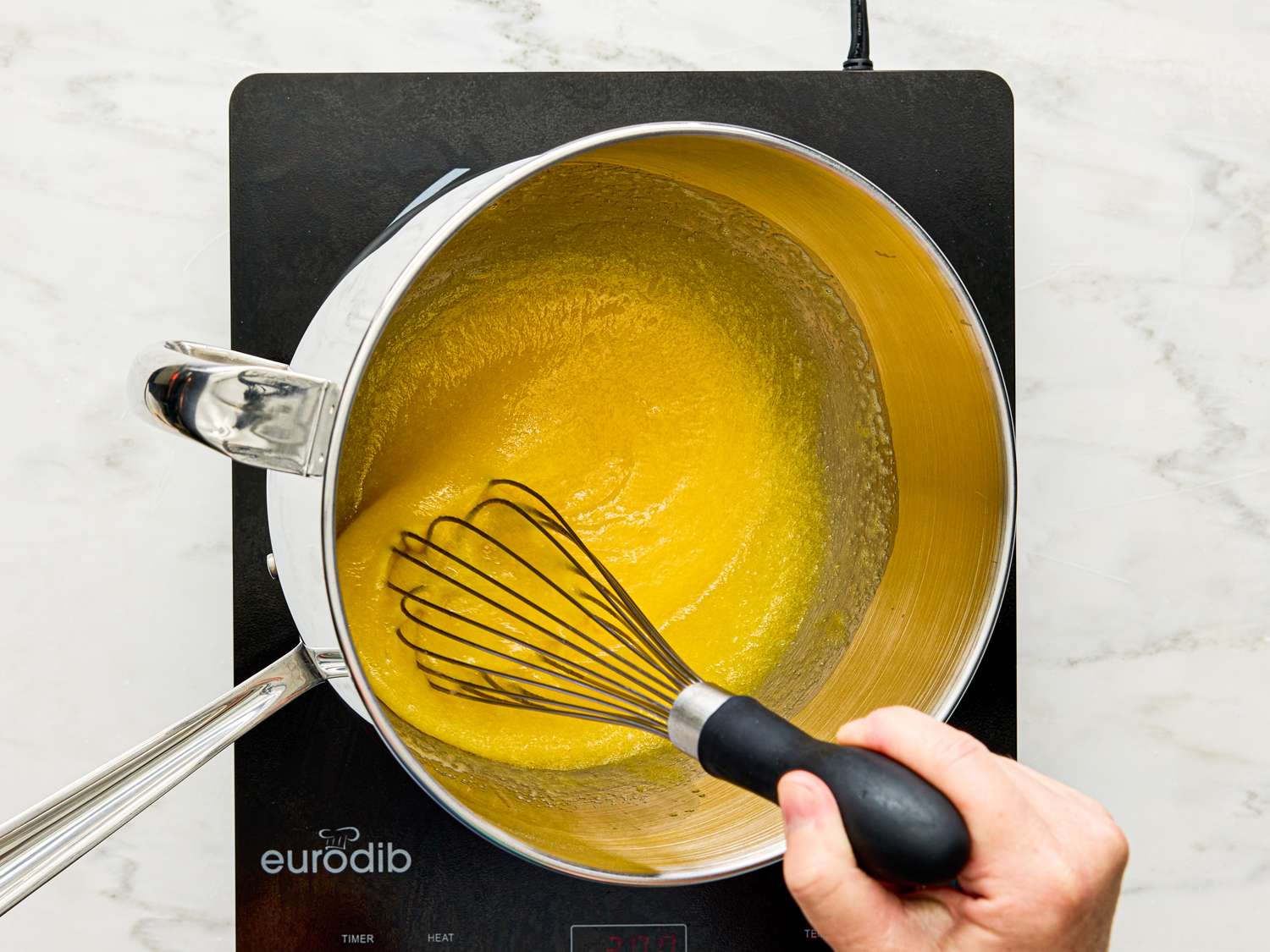 A hand whisking yellow mixture in a saucepan on an induction cooktop