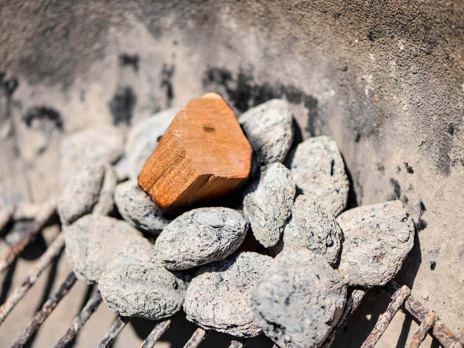 A small pile of ashy charcoal briquettes are placed in a grill. A chunk of smoking wood is nestled atop the briquettes.