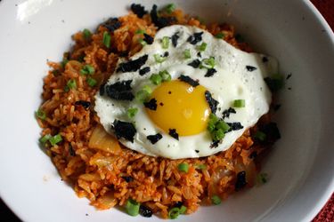 A white bowl holding kimchi fried rice. The rice is topped with a fried egg and sprinkled with dried seaweed.