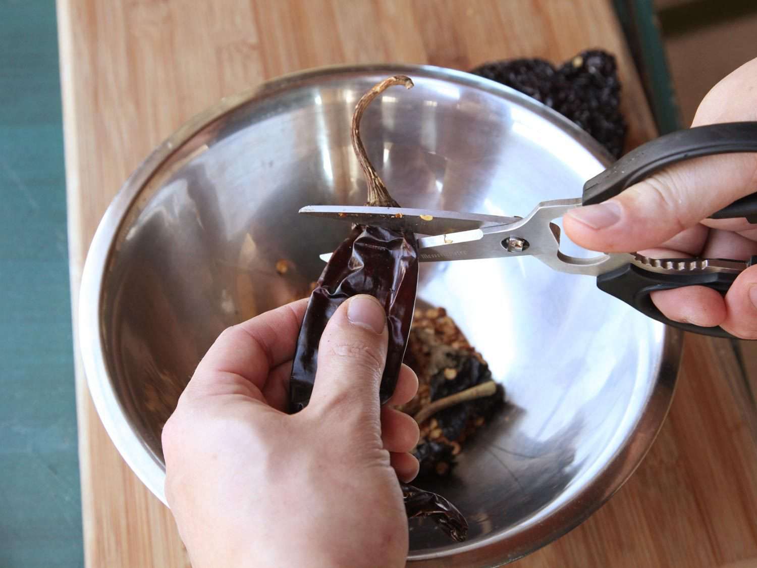 Snipping off the stem of a dried chile with a set of shears, over a metal bowl of other chiles