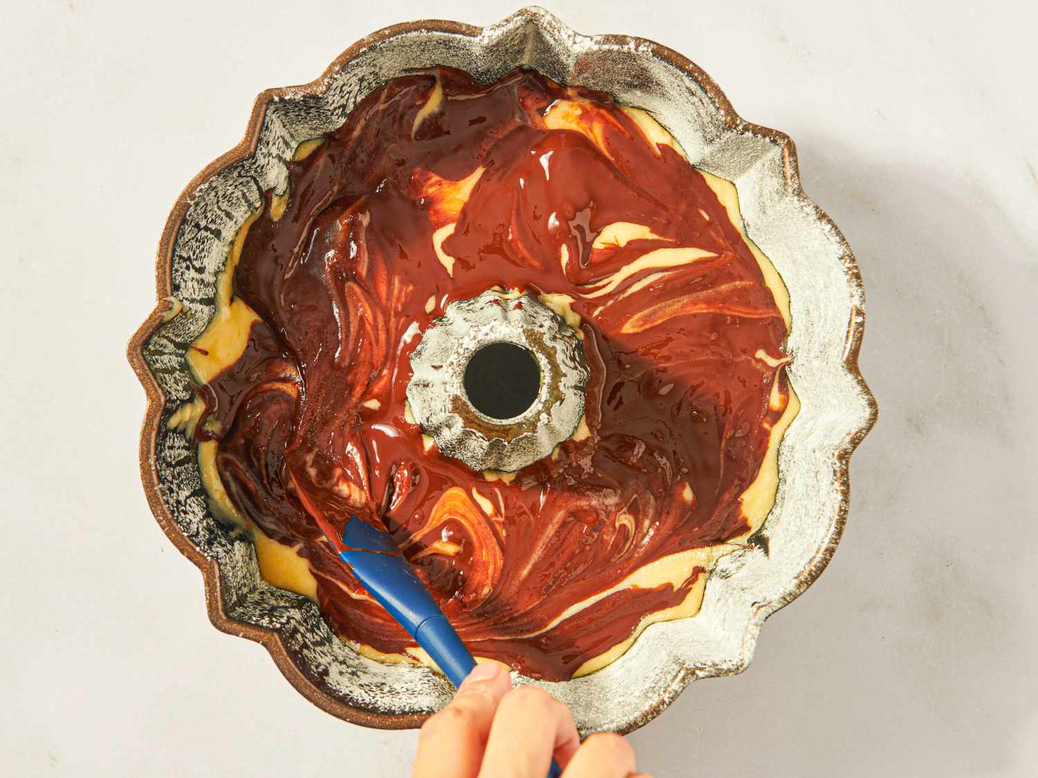 A hand using a spatula to swirl chocolate and vanilla cake batter in a bundt pan