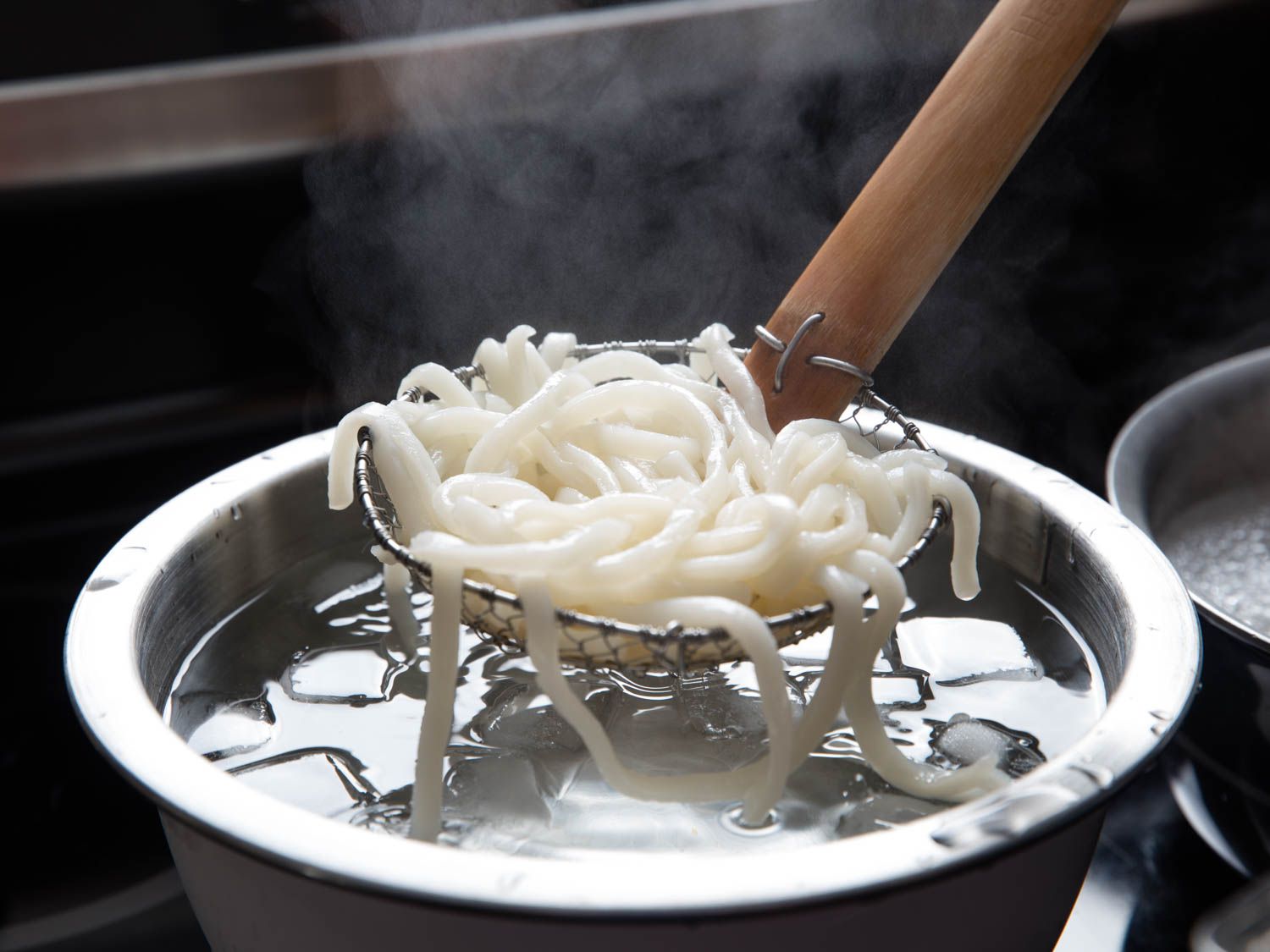 A spider transferring cooked udon noodles to a bowl of ice water.