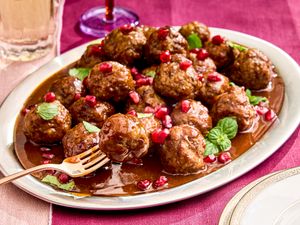 Plate of cocktail meatballs garnished with pomegranate seeds and mint leaves set on a tablecloth with glasses in the background