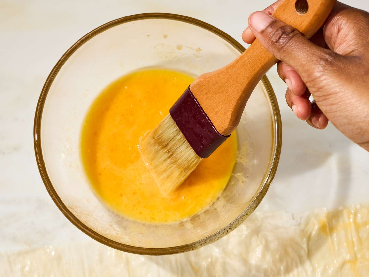 A hand holding a pastry brush over a bowl with an egg wash mixture
