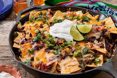 A skillet of nachos topped with melted cheese, black beans, sour cream, lime wedges, and green onions, placed on a wooden surface