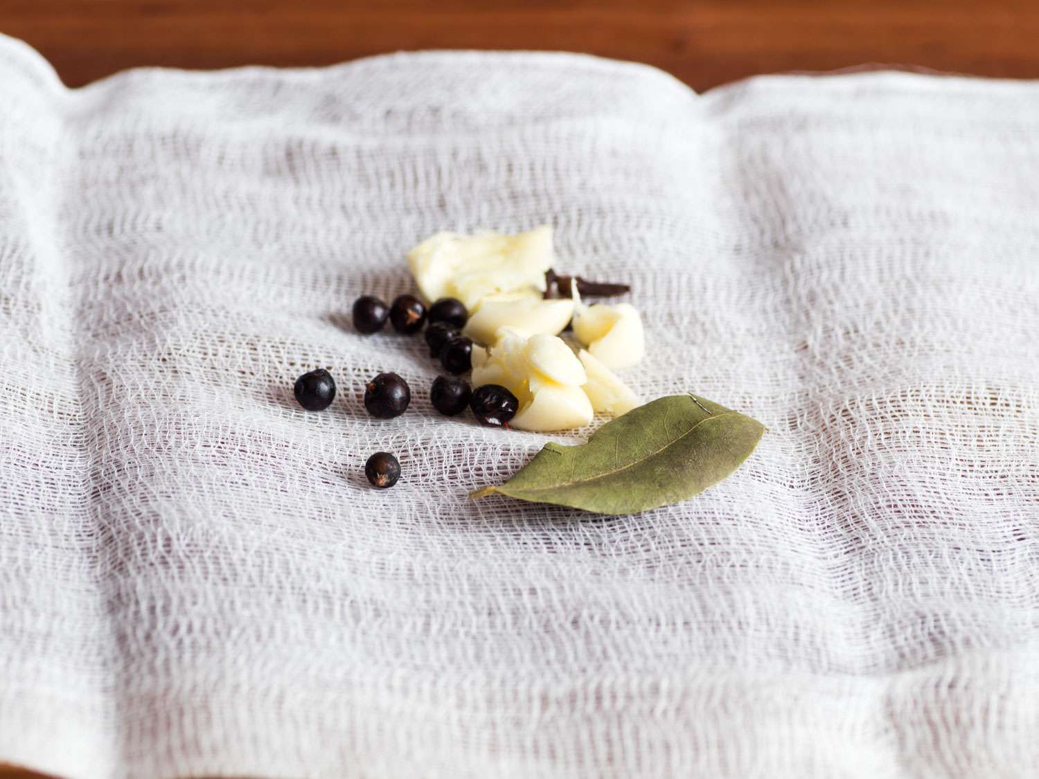Close-up of a square of cheesecloth with smashed garlic, juniper berries, cloves, and a bay leaf laying on top for making choucroute garnie.