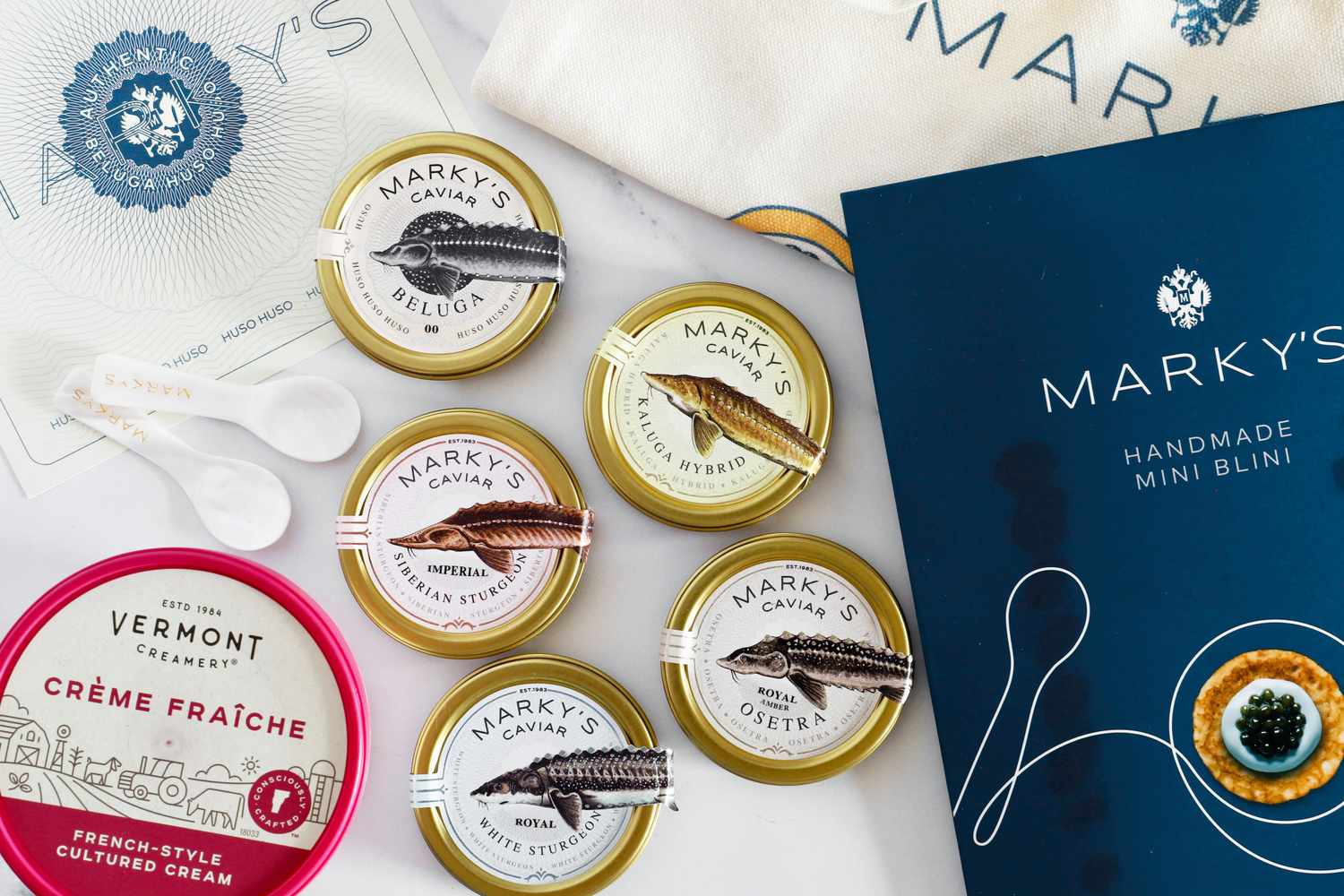 A collection of products from Marky's, including beluga, kaluga, siberian sturgeon, osetra, and white sturgeon caviars, blinis, a tote bag, and crème fraîche