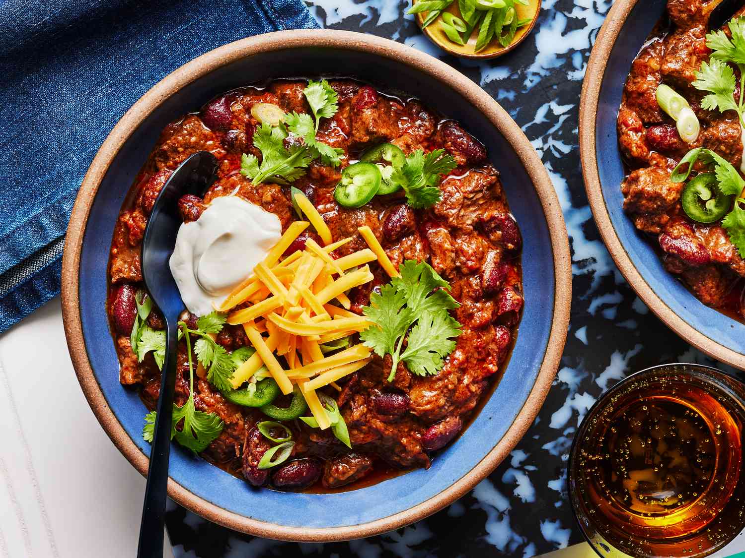 2 bowls of chili in blue bowls, on a blue marble surface, on a white tabletop with small dish of limes, green onions and a drink