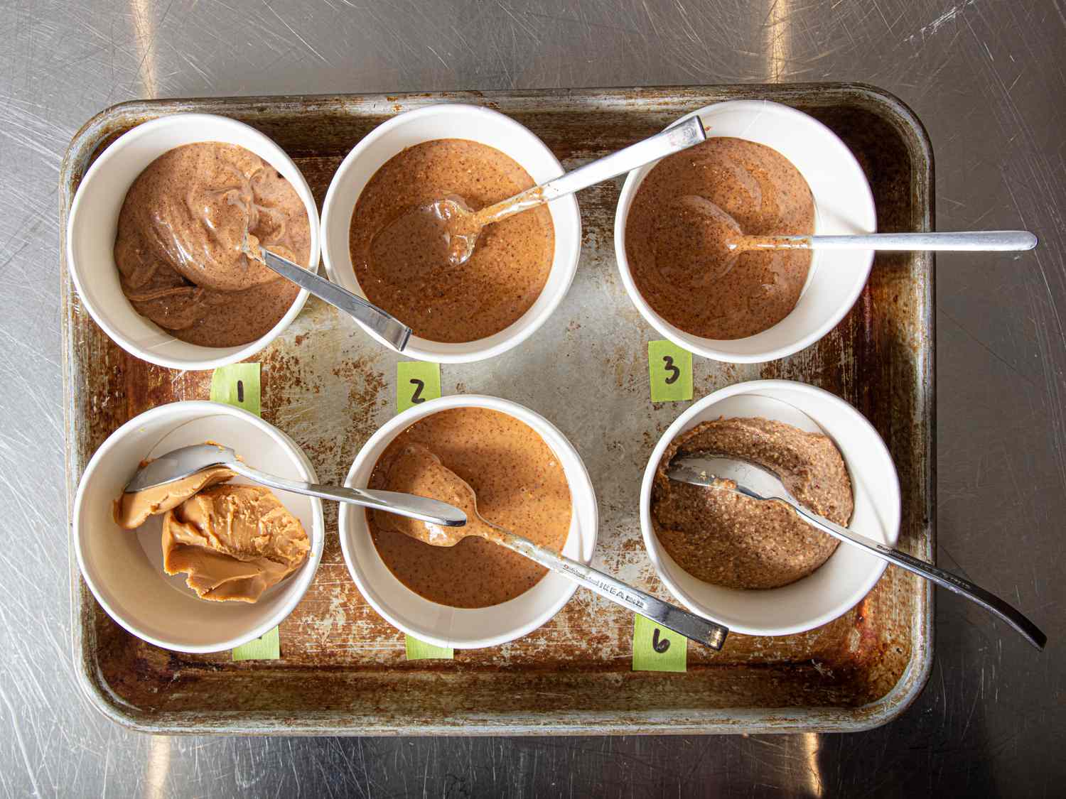 Six bowls labeled with numbers, each containing portions of nut butter and a spoon, arranged on a tray