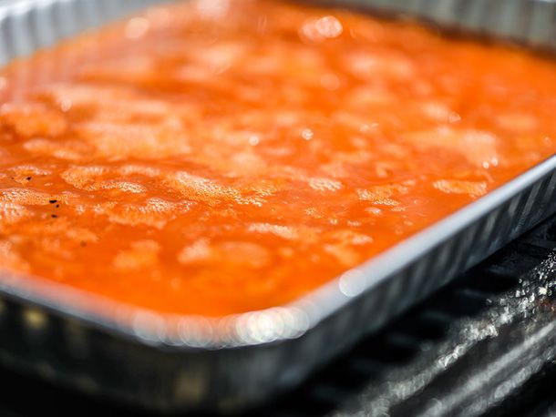 An aluminum roasting pan, sitting on a grill, filled with a bubbling, reddish-orange mixture for poaching hotdogs. 