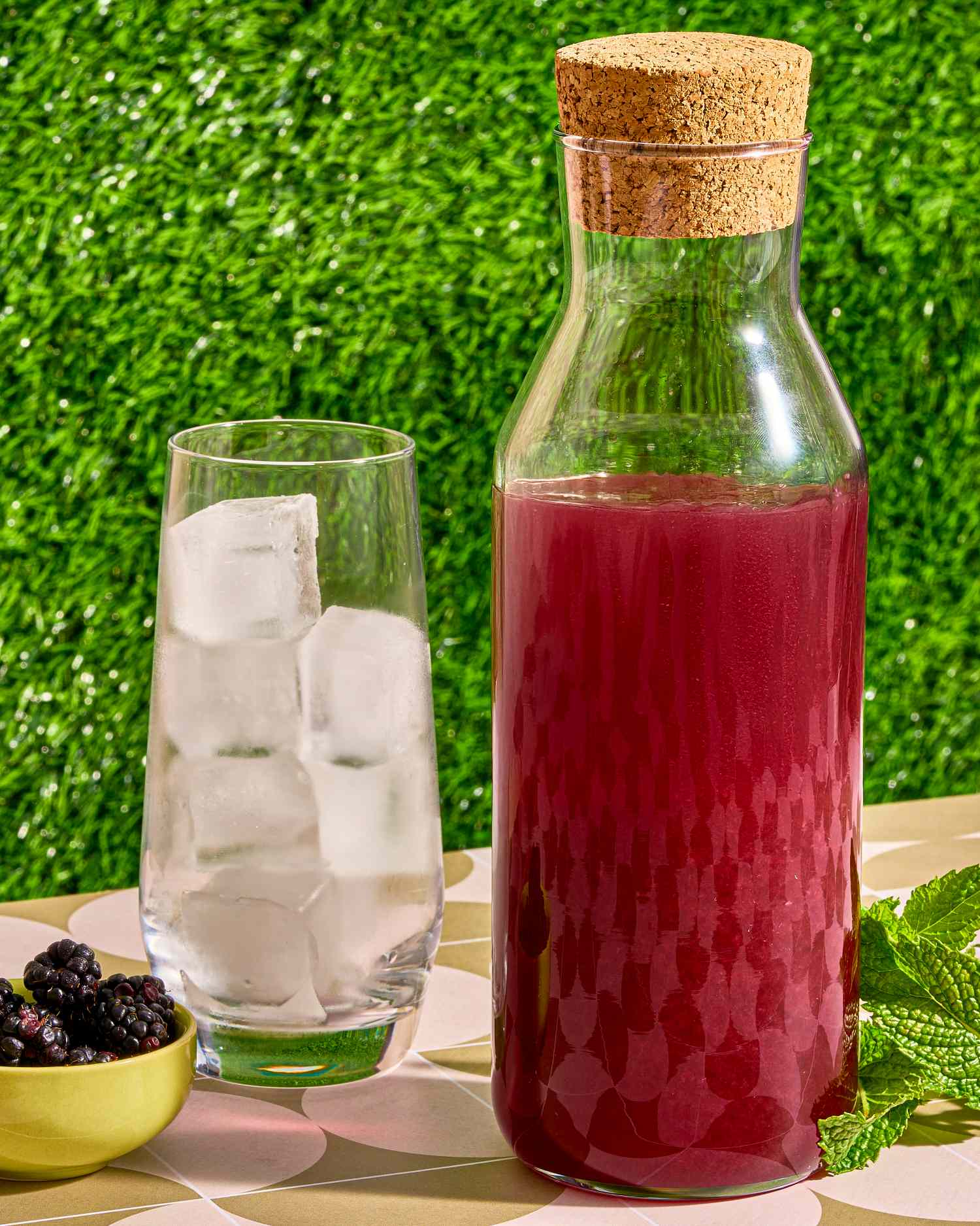 A bottle of blackberry liqueur next to a glass of ice and a bowl of blackberries, set on a table near greenery