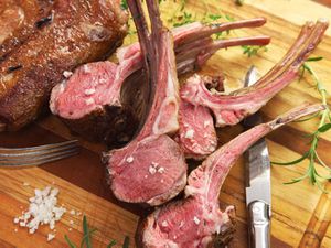 A rack of lamb cooked sous vide and sliced into individual portions, all fanned out on a wooden cutting board with a pile of salt and sprigs of fresh herbs all around.