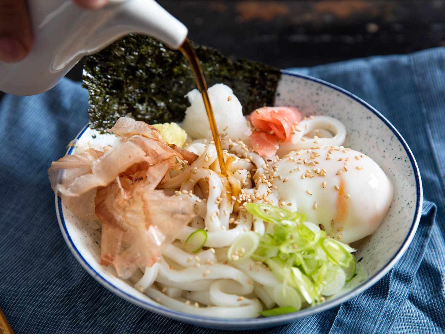 Chilled kaeshi broth being poured from the spout of a ceramic vessel into a fully-loaded bowl of bukkake udon.