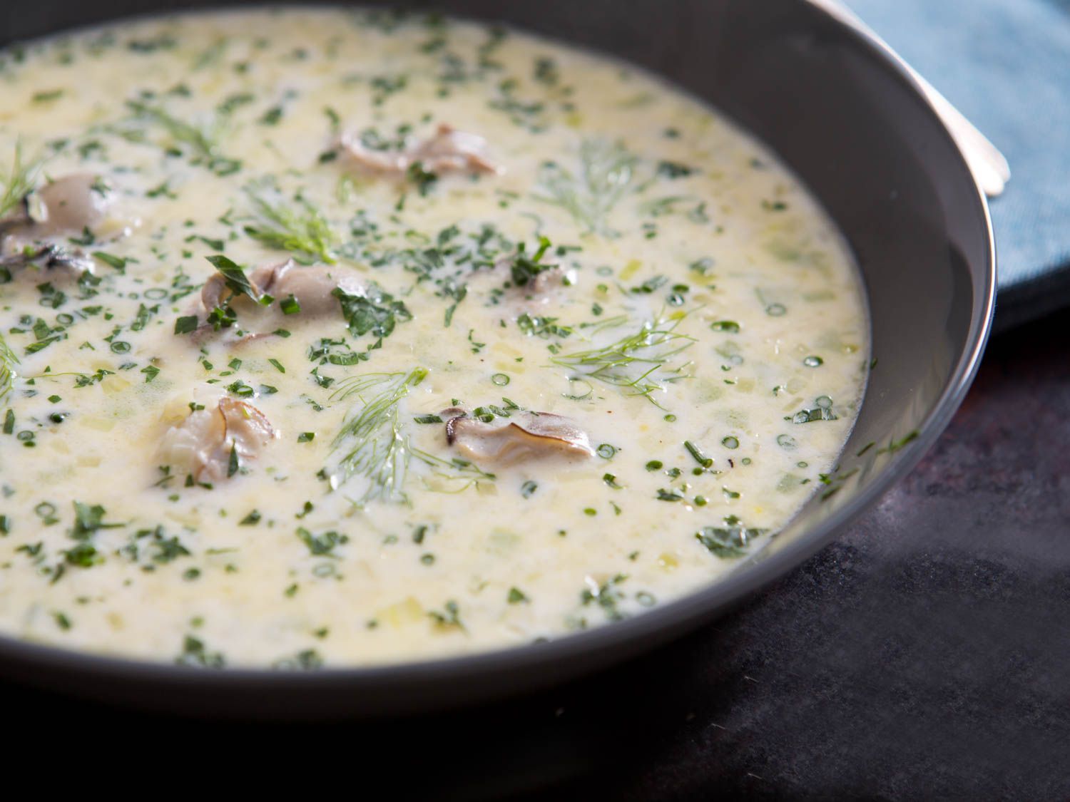 Closeup of oyster stew served in a black bowl, garnished with minced chive, parsley, and fennel fronds.