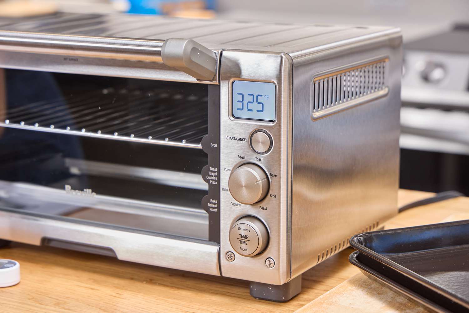 A breville compact toaster oven set to 325 degrees Fahrenheit displayed on a wooden countertop, a baking tray nearby