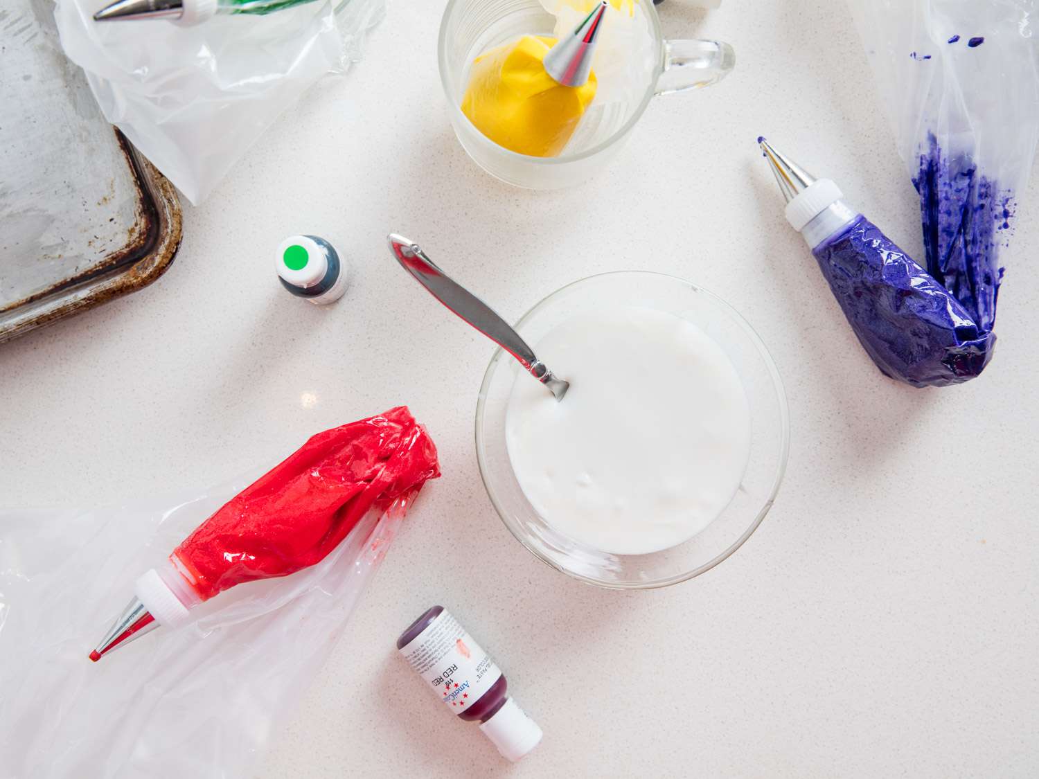 Tools for frosting cookies with colorful royal icing: bowl of plain white royal icing, bottles of gel paste food dye, and piping bags of colored icing in red, yellow, and blue.